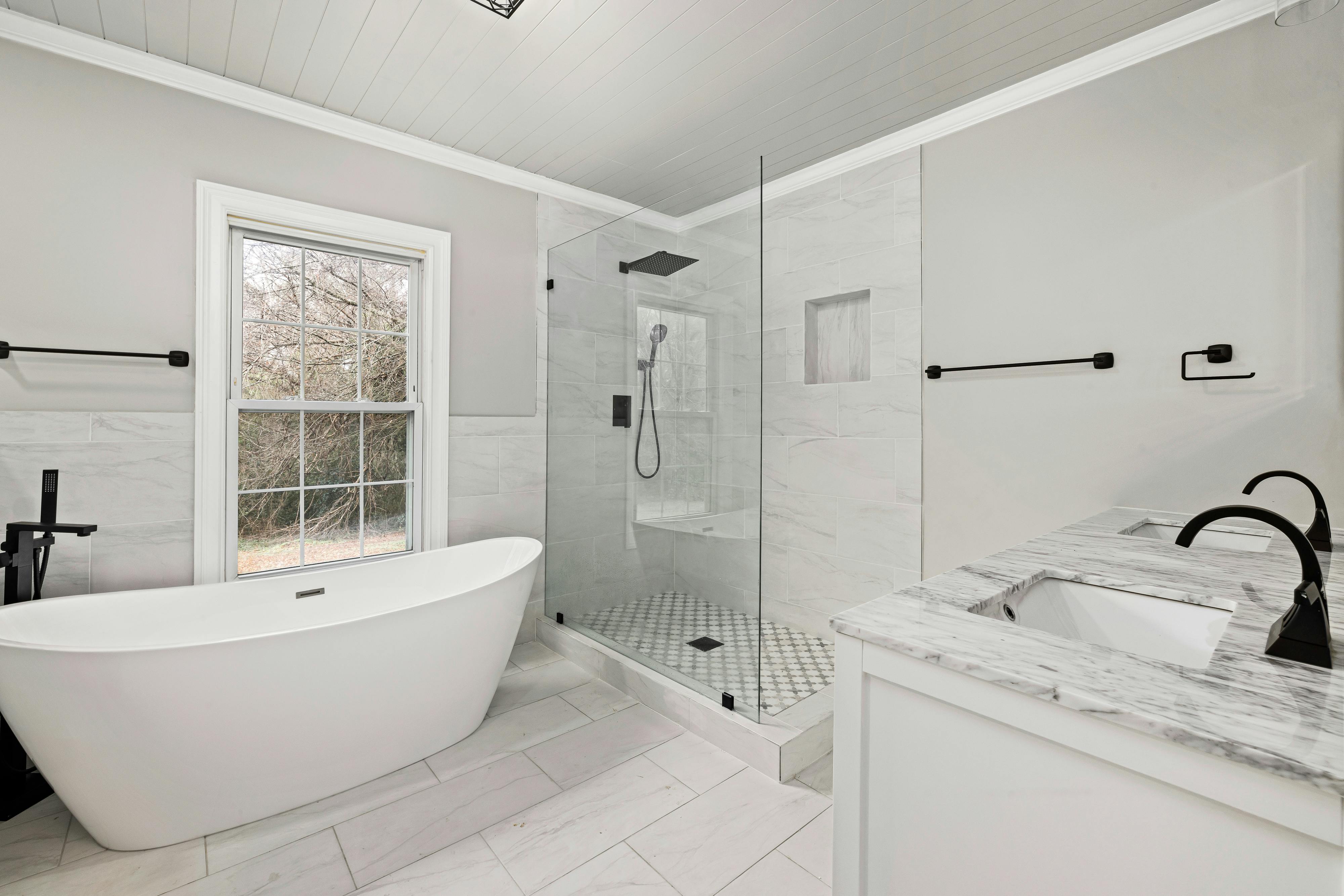 Modern bathroom with a white freestanding bathtub, glass-enclosed shower with black fixtures, and dual sink vanity with marble countertop.
