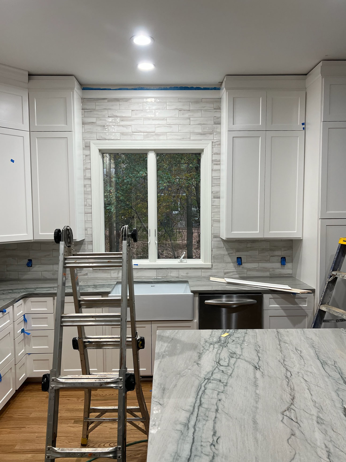 Modern white kitchen under renovation with marble countertops, a farmhouse sink under a window, and subway tile backsplash.