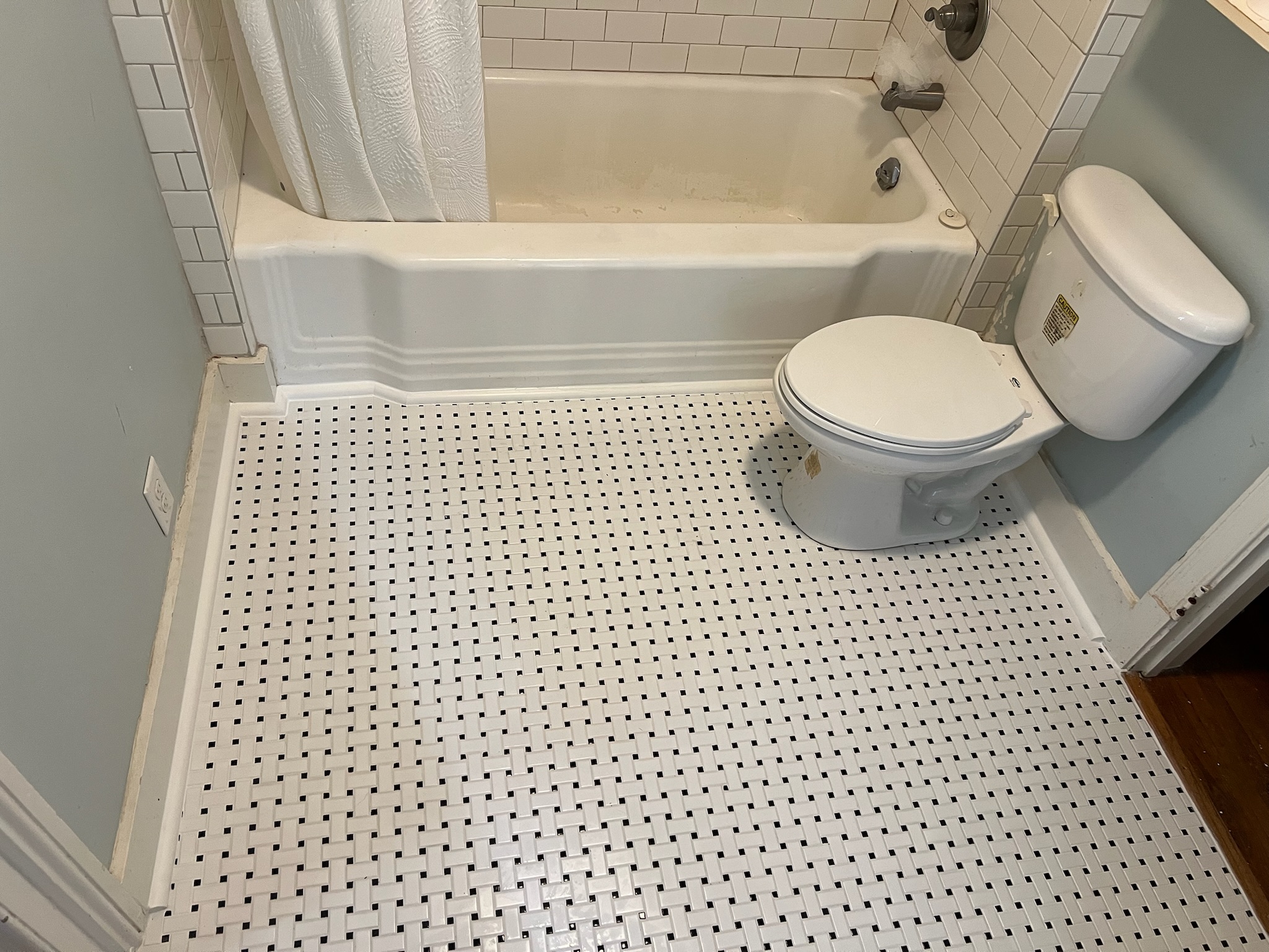 Bathroom corner with a white toilet, bathtub with shower curtain, and a black and white basketweave tiled floor.