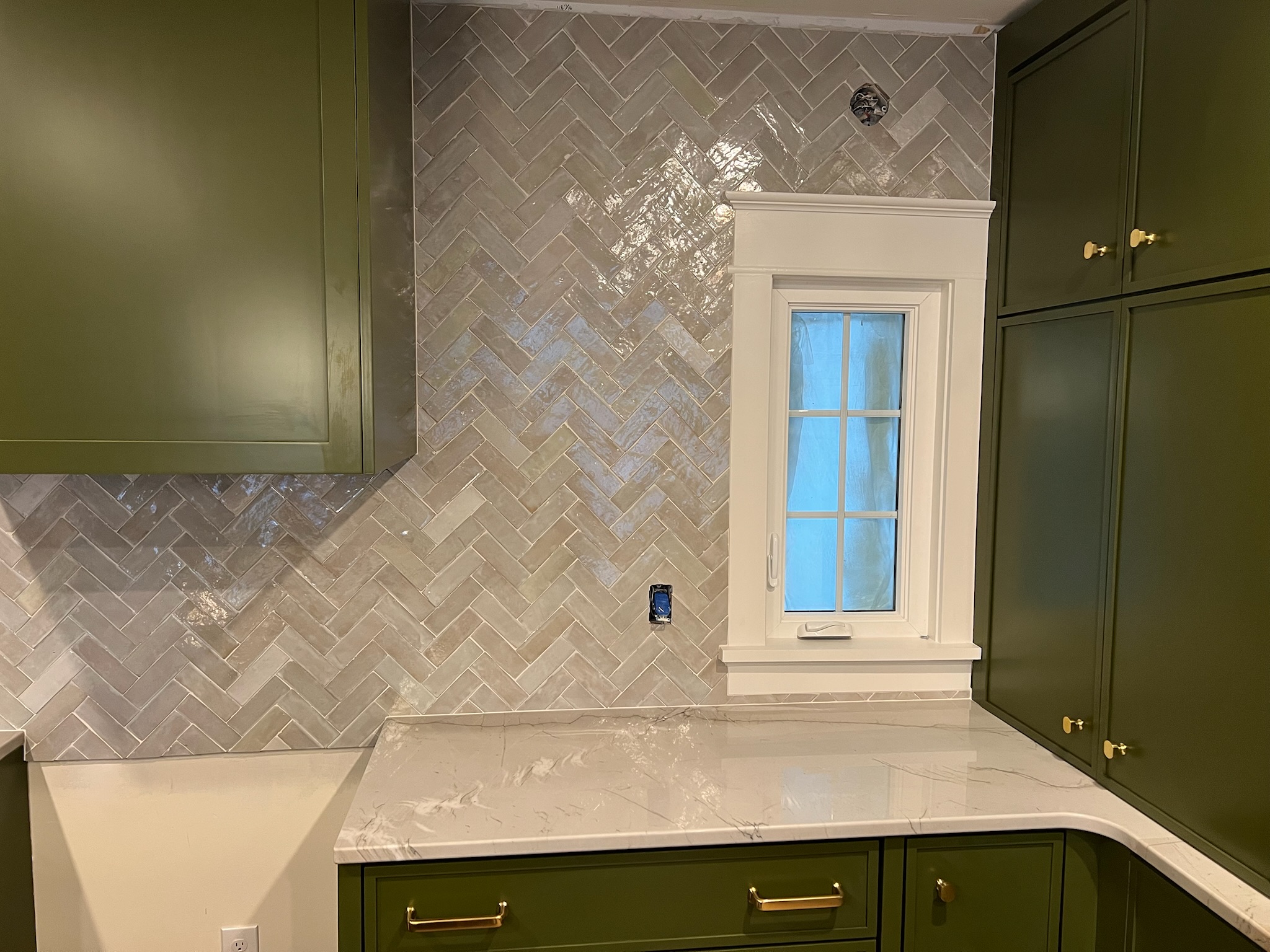 Kitchen corner with green cabinets, white marble countertop, small window with white trim, and glossy beige herringbone tile backsplash.
