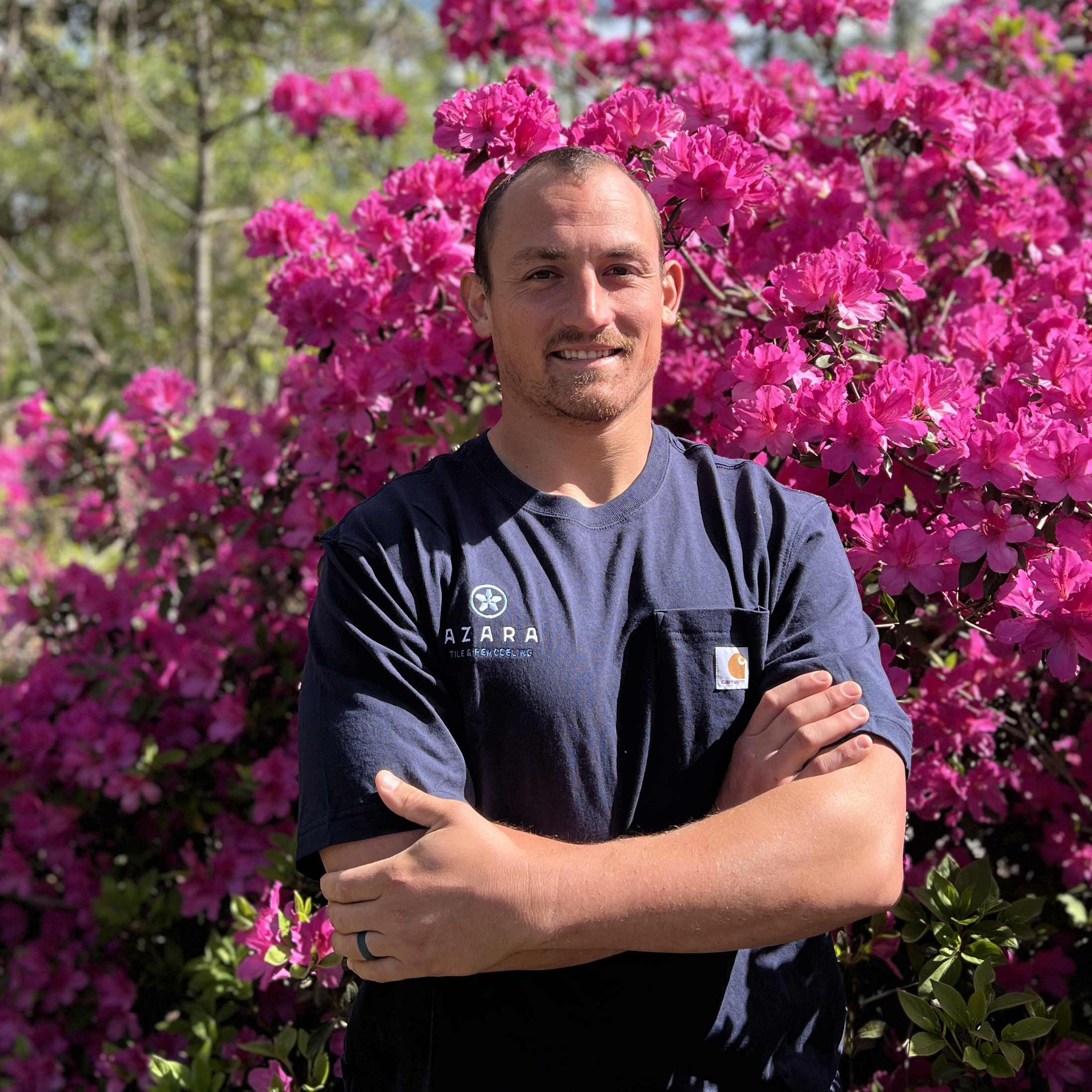 Man wearing a navy blue Azara Tile & Remodeling t-shirt standing with arms crossed in front of bright pink flowering bushes.