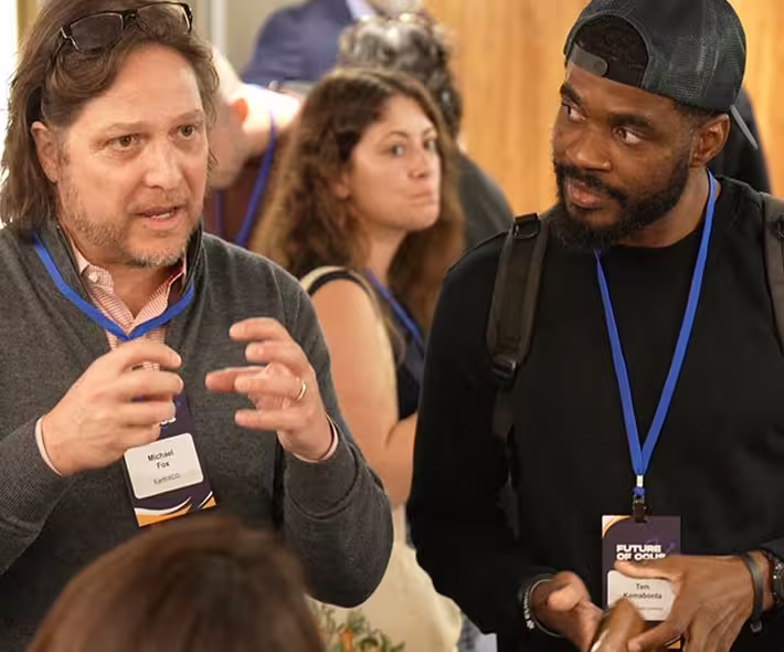 Two men wearing name tags engaged in conversation at an event, with a woman and other attendees blurred in the background.