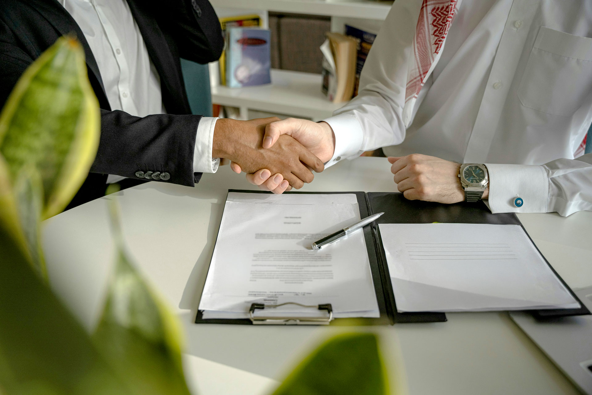 Two men shaking hands over a white table with documents and a pen, one wearing a black suit and the other a white traditional garment with a watch.