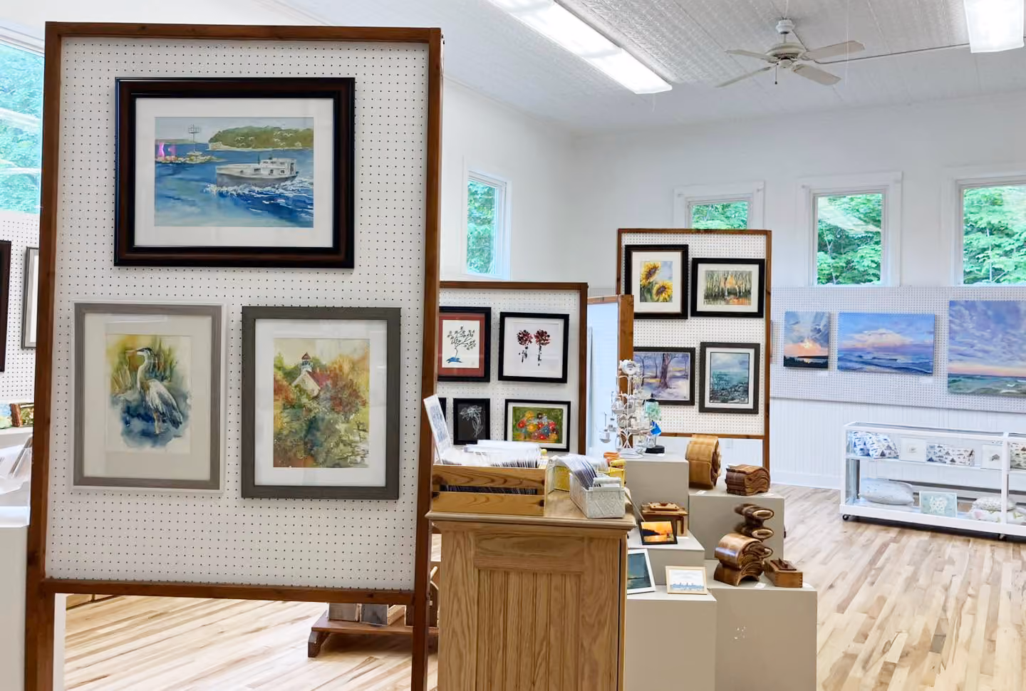 Art gallery room with watercolor paintings and wooden crafts displayed on panels and pedestals.