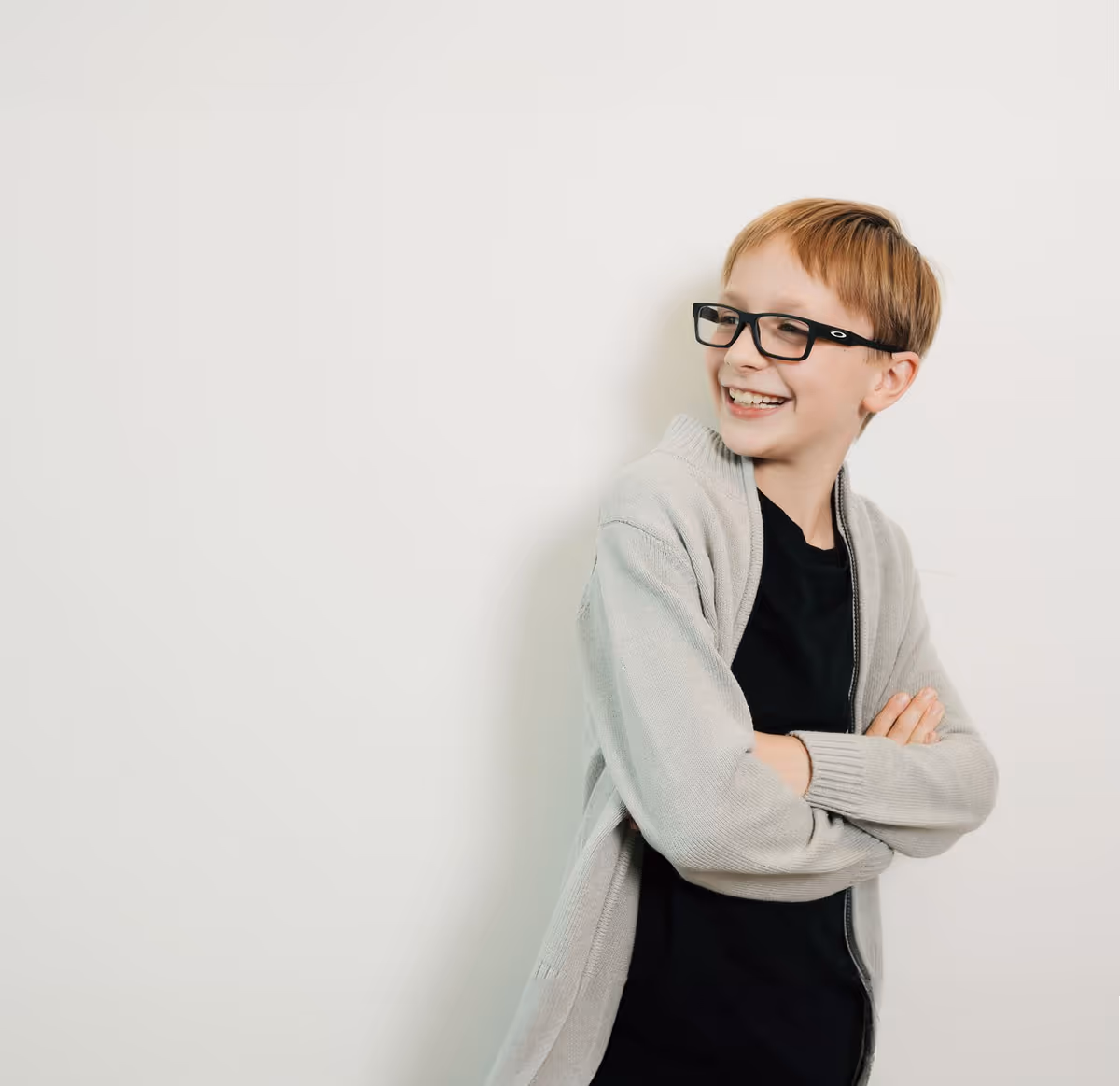 Smiling young boy with glasses wearing a gray cardigan and black shirt against a white wall.