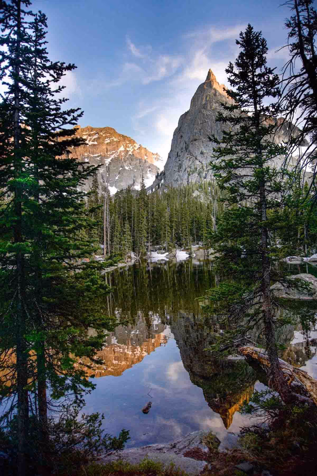 Mountain peak reflected in a clear lake surrounded by pine trees under a blue sky.