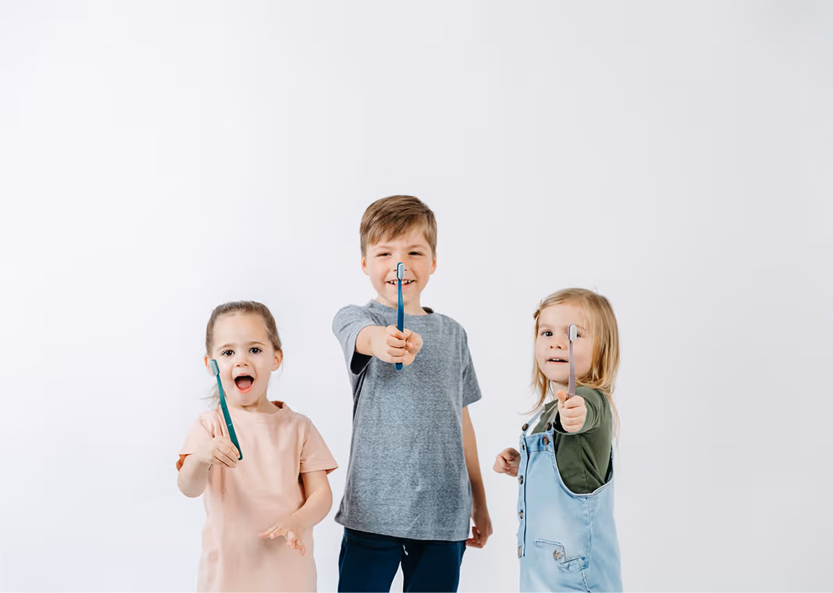 Three children smiling and holding toothbrushes toward the camera against a white background.