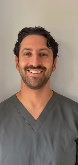 Smiling man with curly dark hair wearing gray scrubs against a plain light background.
