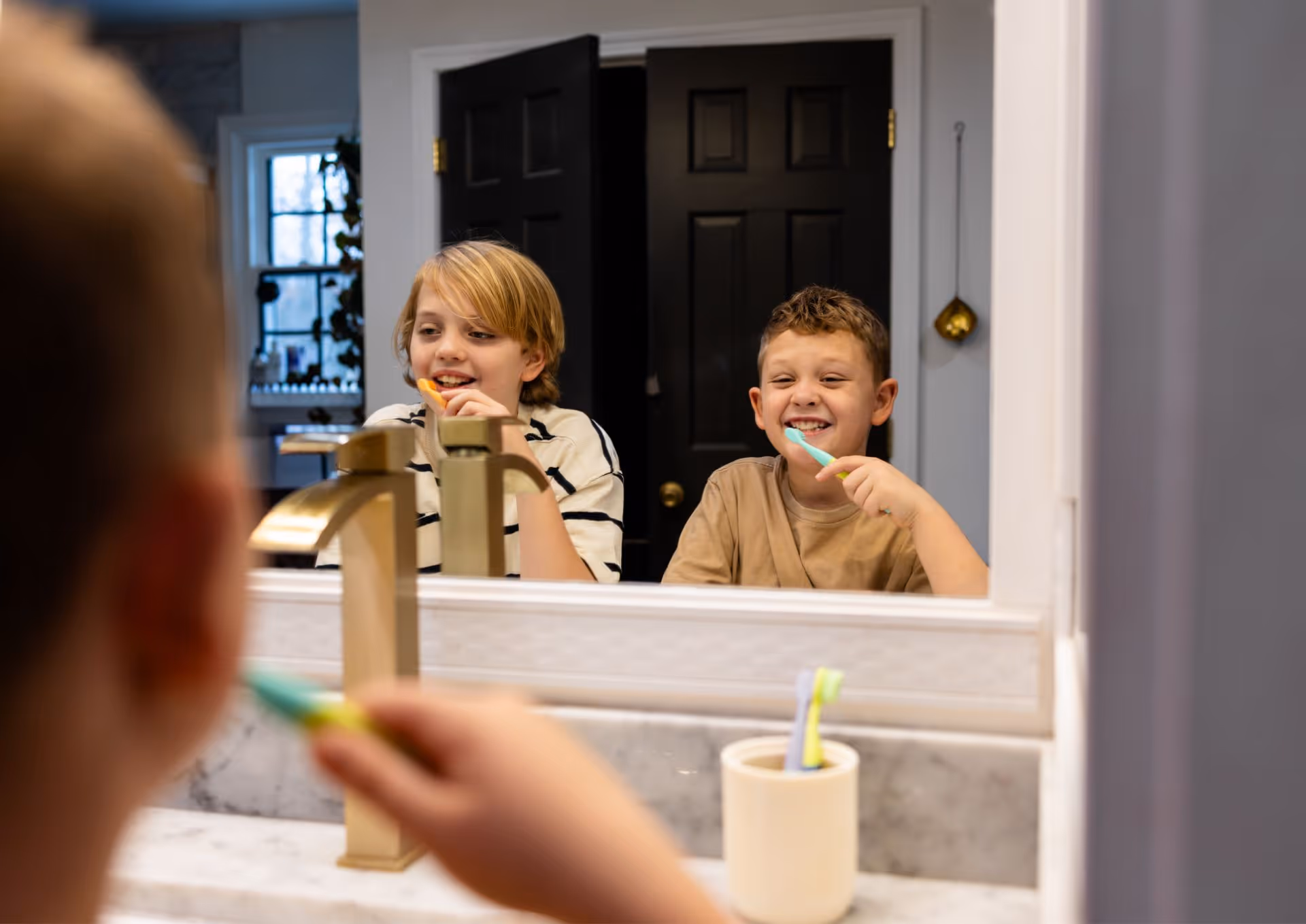Two boys smiling while brushing their teeth together in front of a bathroom mirror.