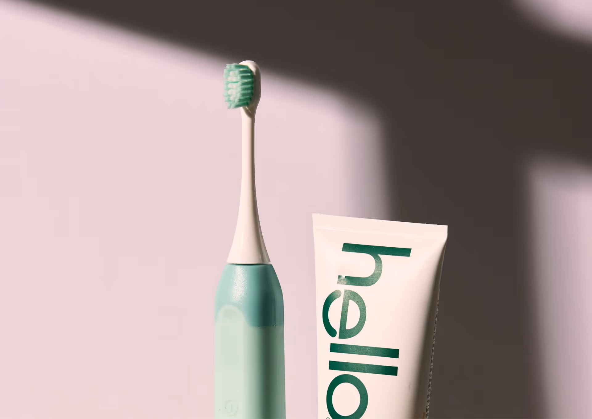 Electric toothbrush with green bristles next to a white tube of Hello toothpaste with green text.