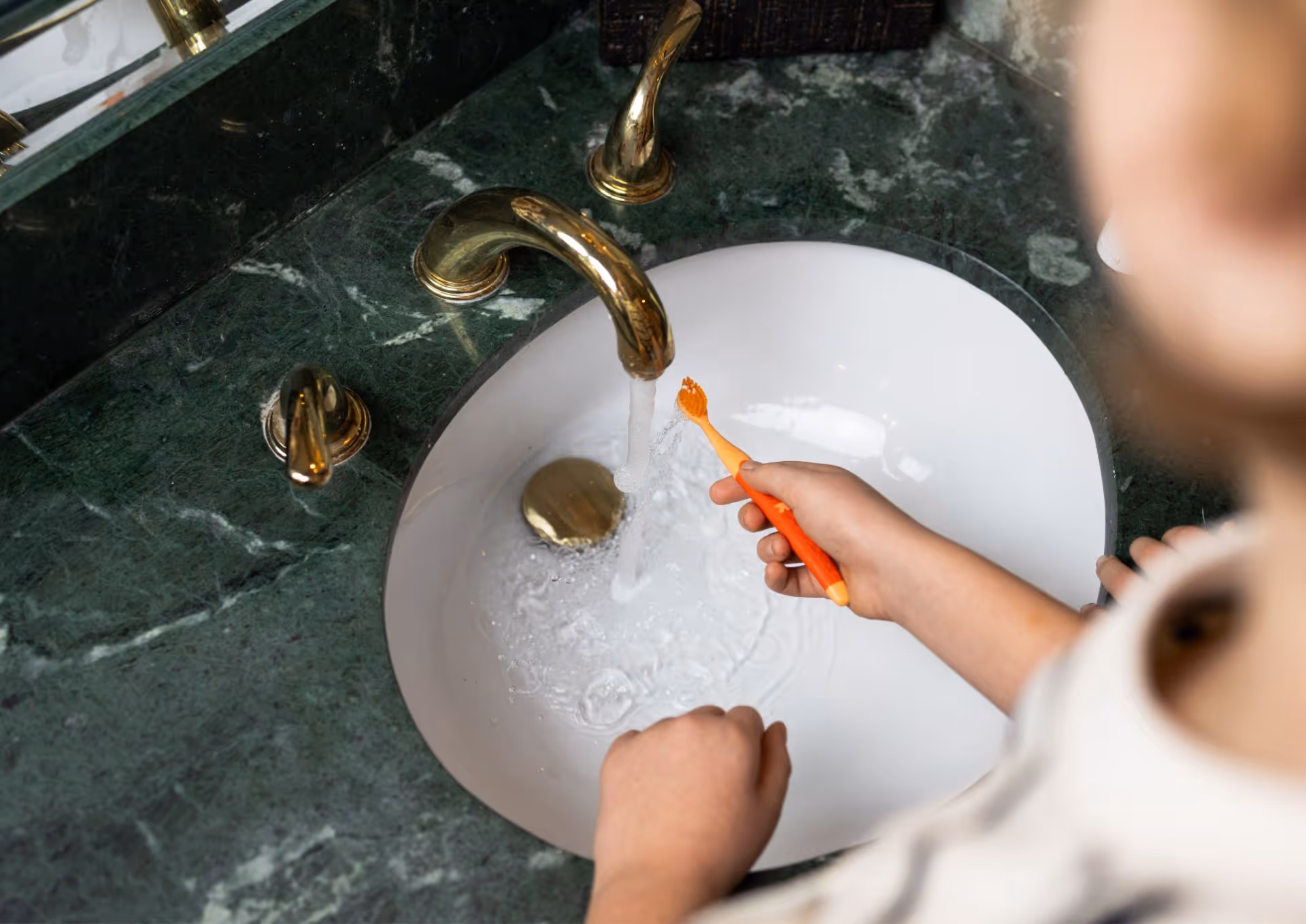 Child holding orange toothbrush under running water in a white sink with gold fixtures.