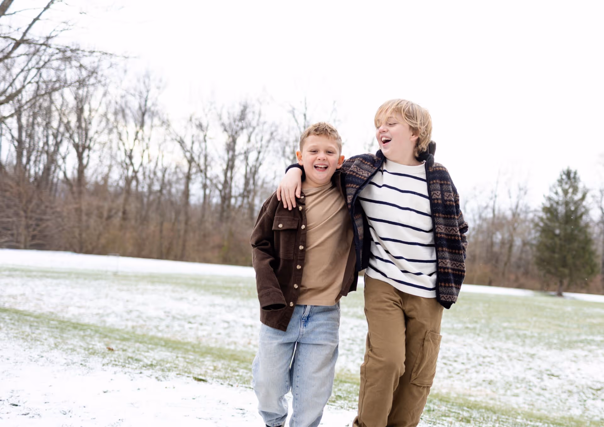 Two boys walking arm in arm on snow-dusted grass with bare trees in background.