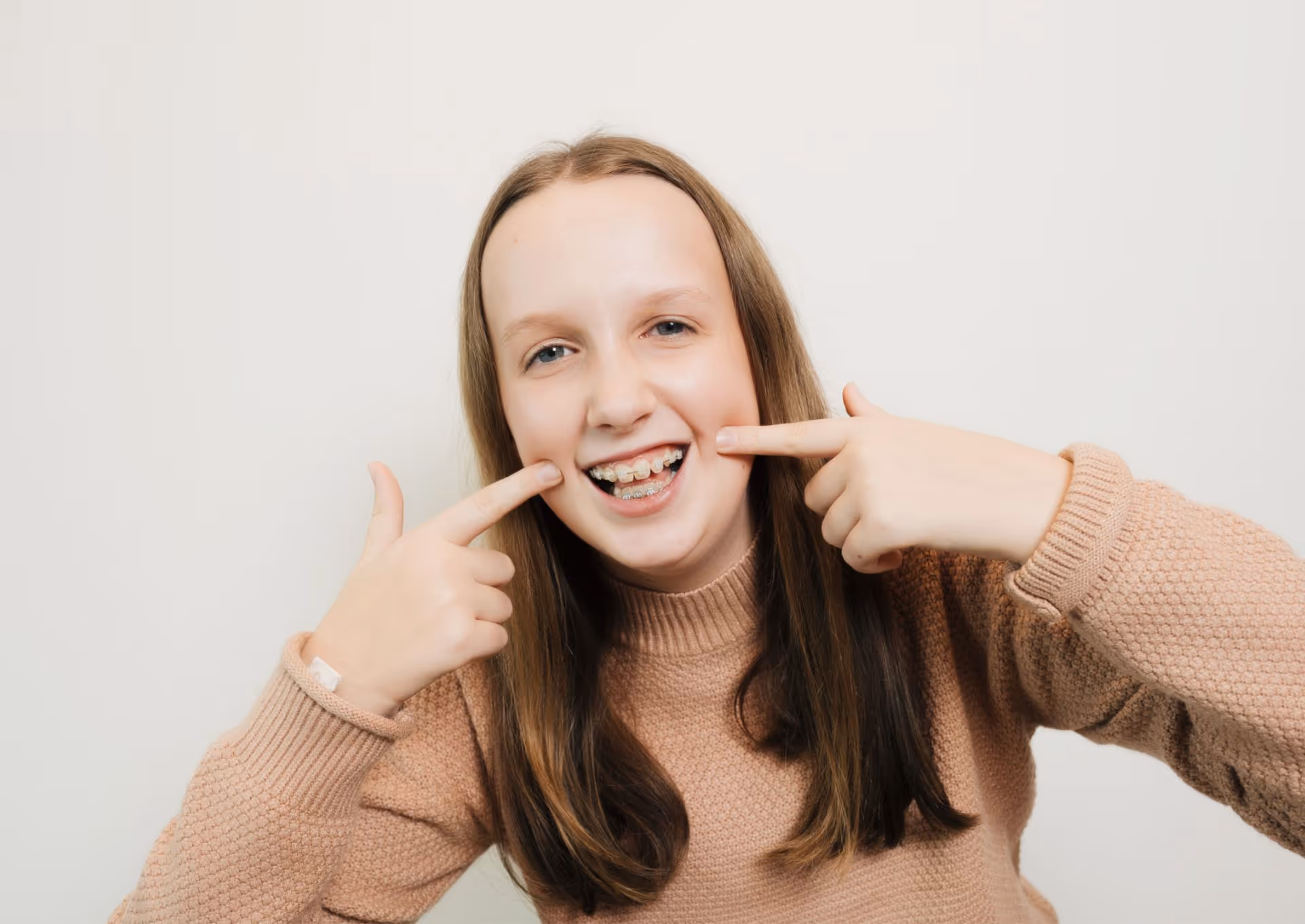 Smiling girl pointing at her braces on her teeth wearing a tan sweater against a plain background.