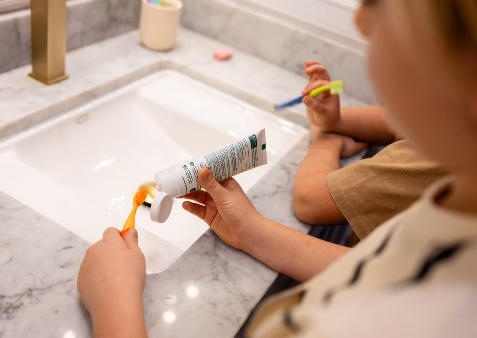 Two children at a bathroom sink, one squeezing toothpaste onto an orange toothbrush.