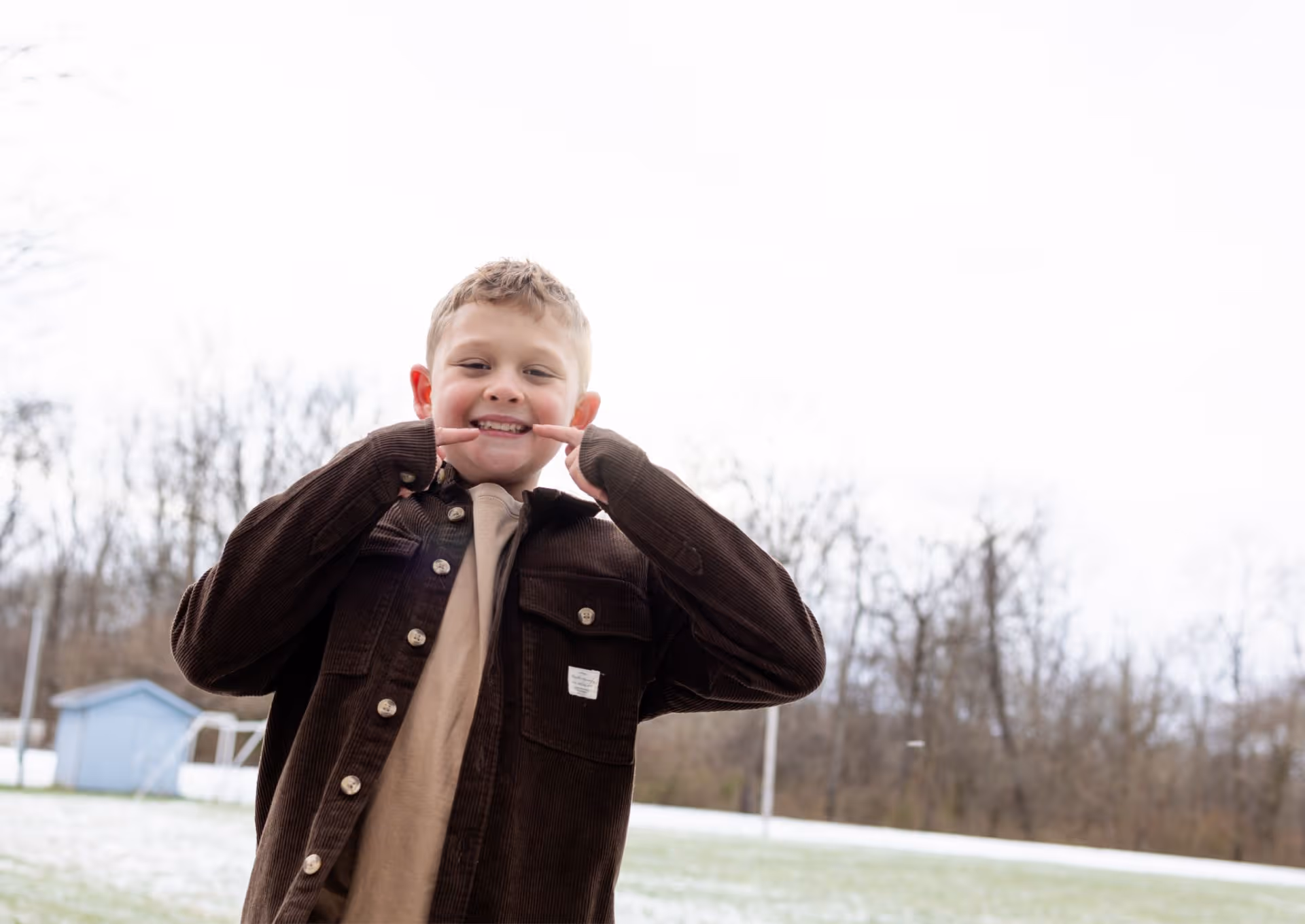 Boy outside in a brown jacket smiling and pointing to his teeth with his fingers.