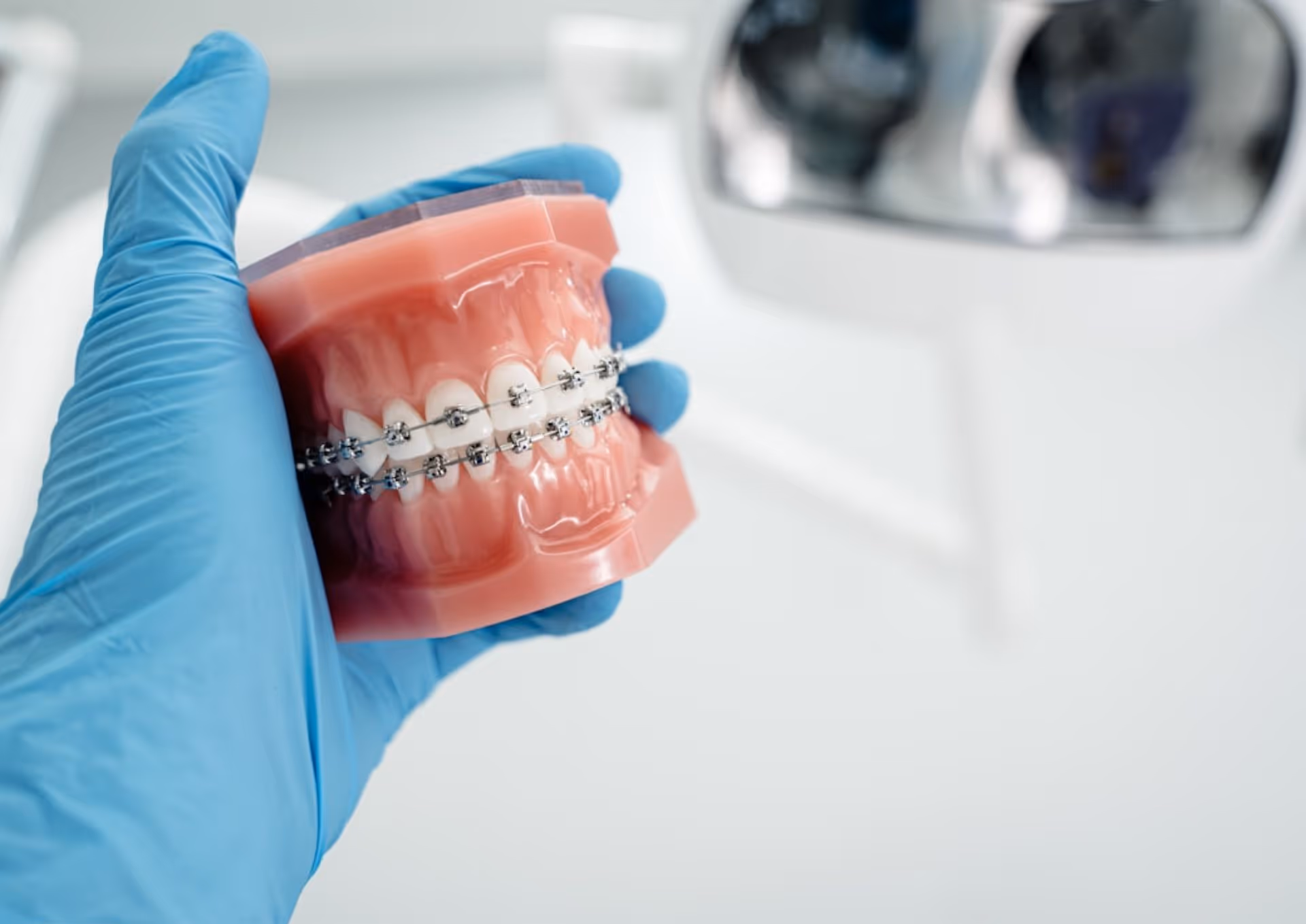 Gloved hand holding dental model with braces on upper and lower teeth in dental clinic.