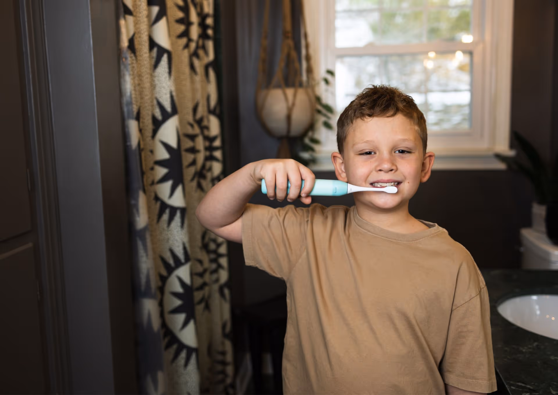 Young boy in beige shirt brushing his teeth with an electric toothbrush in a bathroom.