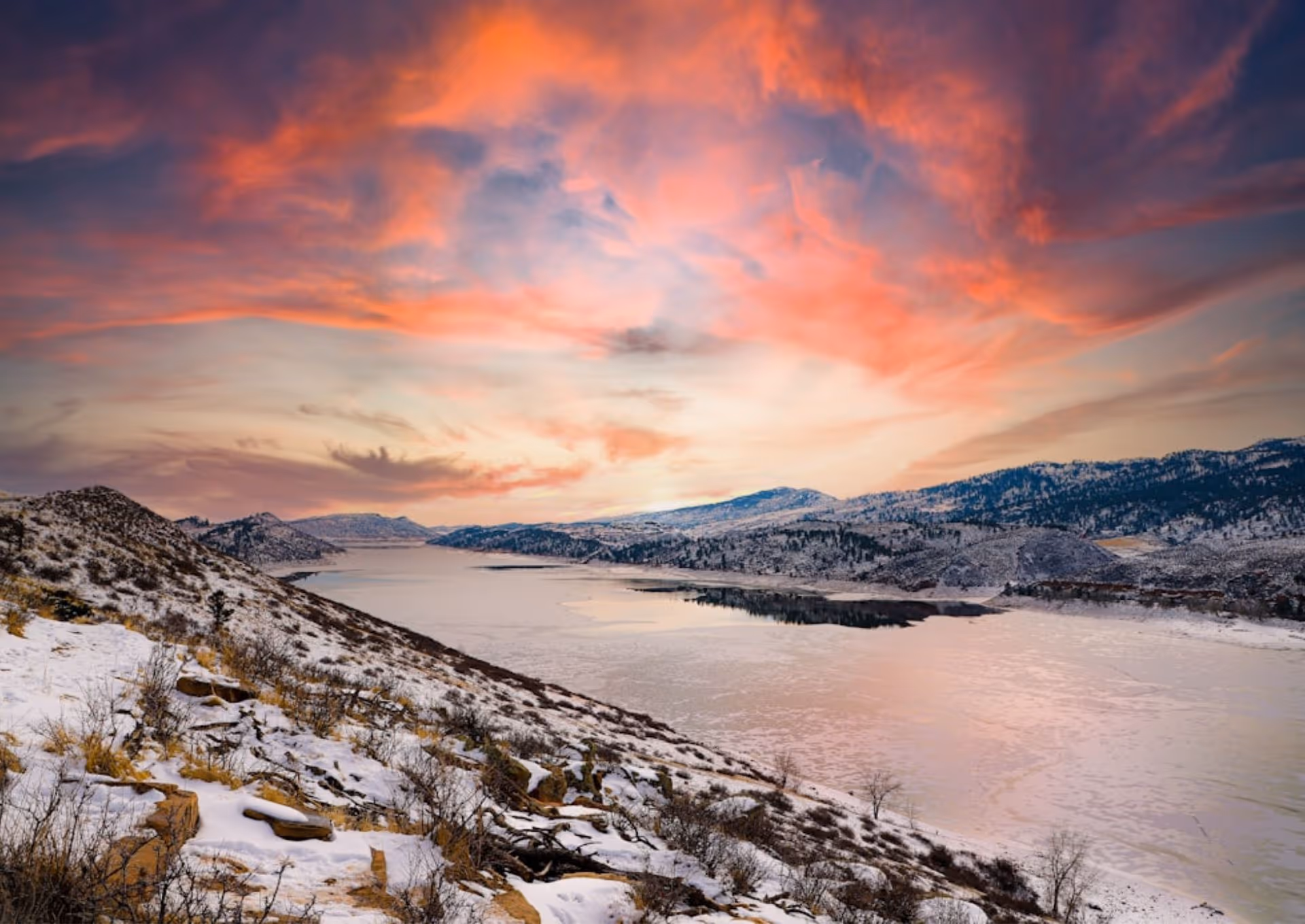 Snow-covered hillside overlooking a frozen lake with mountains under a colorful sunset sky.