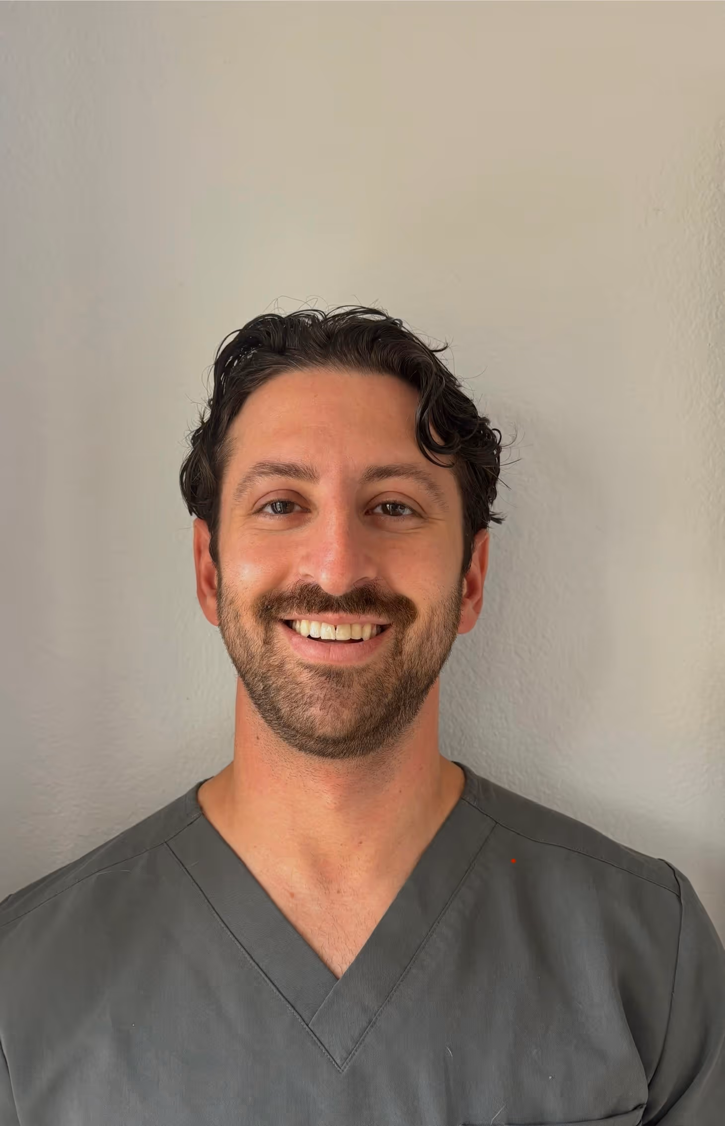 Smiling man with short dark hair and beard wearing a gray V-neck scrub top against a plain wall.