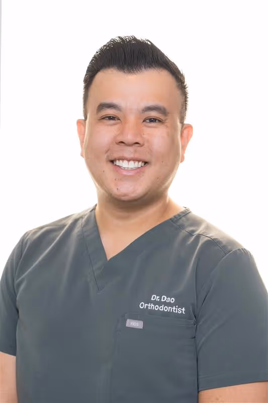 Smiling male orthodontist wearing gray scrubs with 'Dr. Dao Orthodontist' embroidered on shirt.