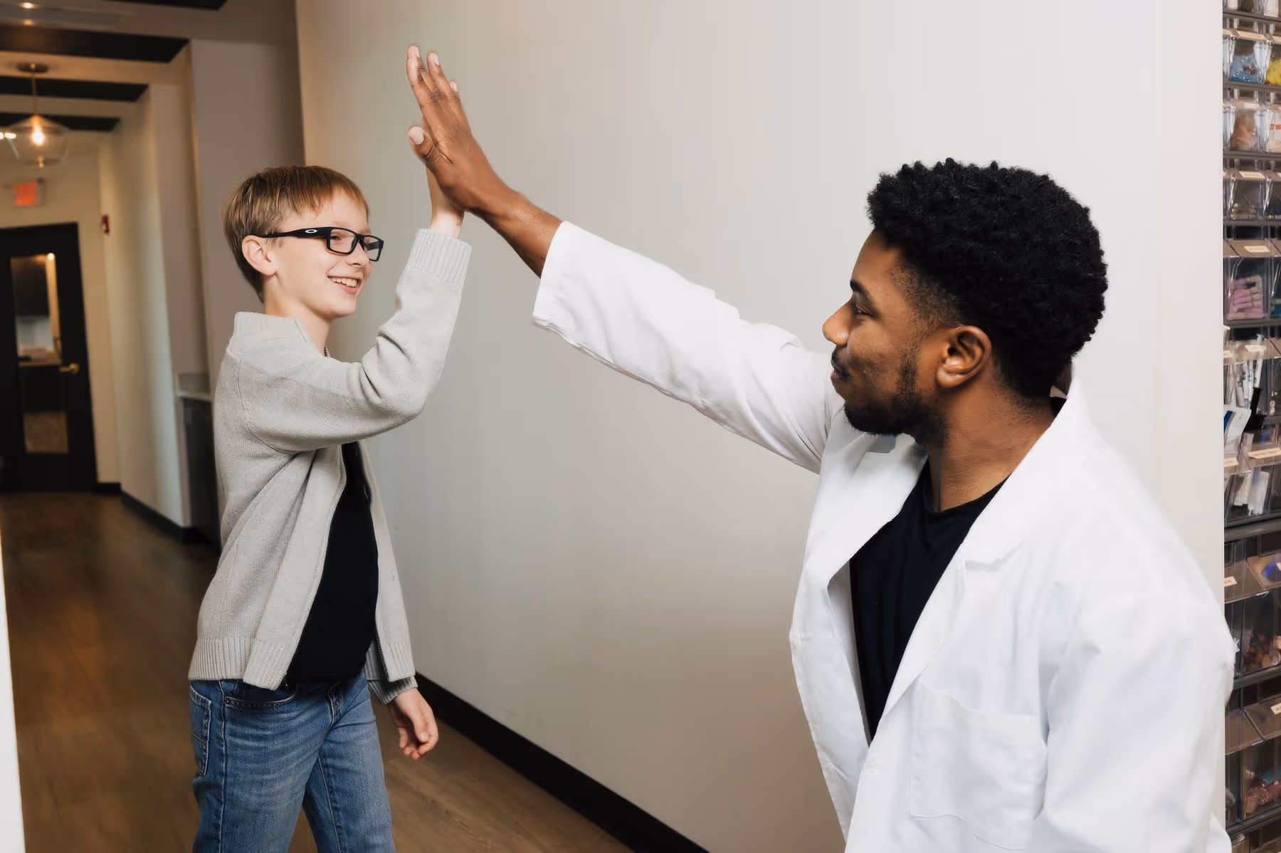 Boy wearing glasses and sweater high-fives a man in a white lab coat in a hallway.