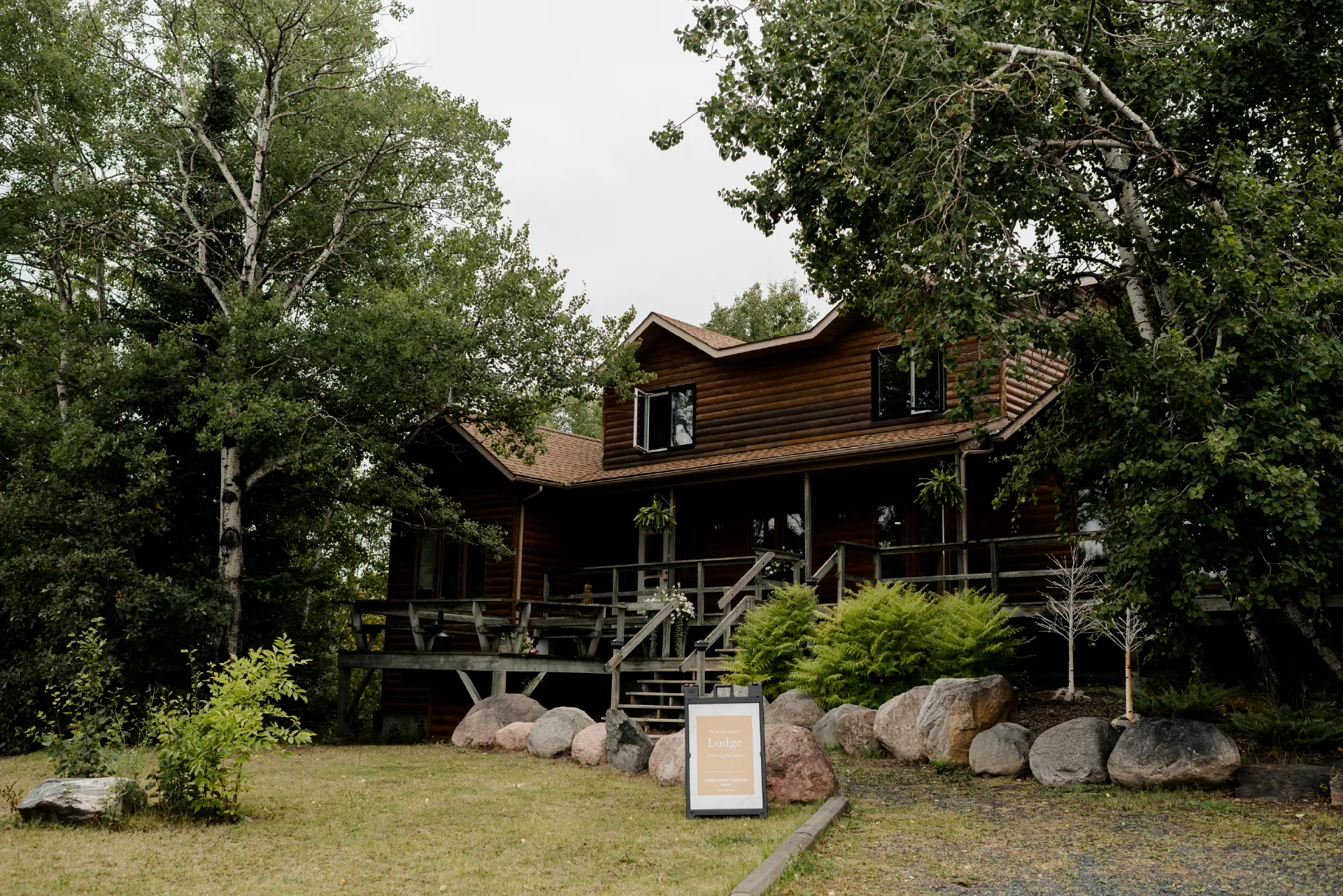 Exterior of a log lodge surrounded by trees, boulders, ferns and a sign for The Lodge at Perch Bay.