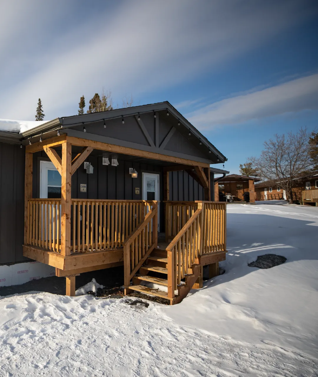 Dark grey cabin with a wooden front porch and stairs surrounded by snow.