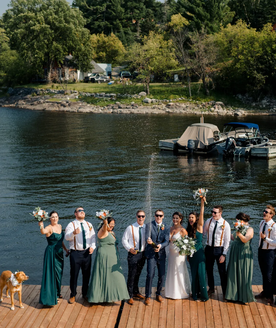 Wedding party celebrating on a wooden dock beside the river, with boats and shoreline in the background.
