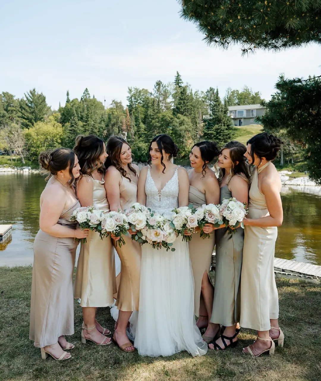 Bride and bridesmaids holding bouquets together on the grass beside the river.