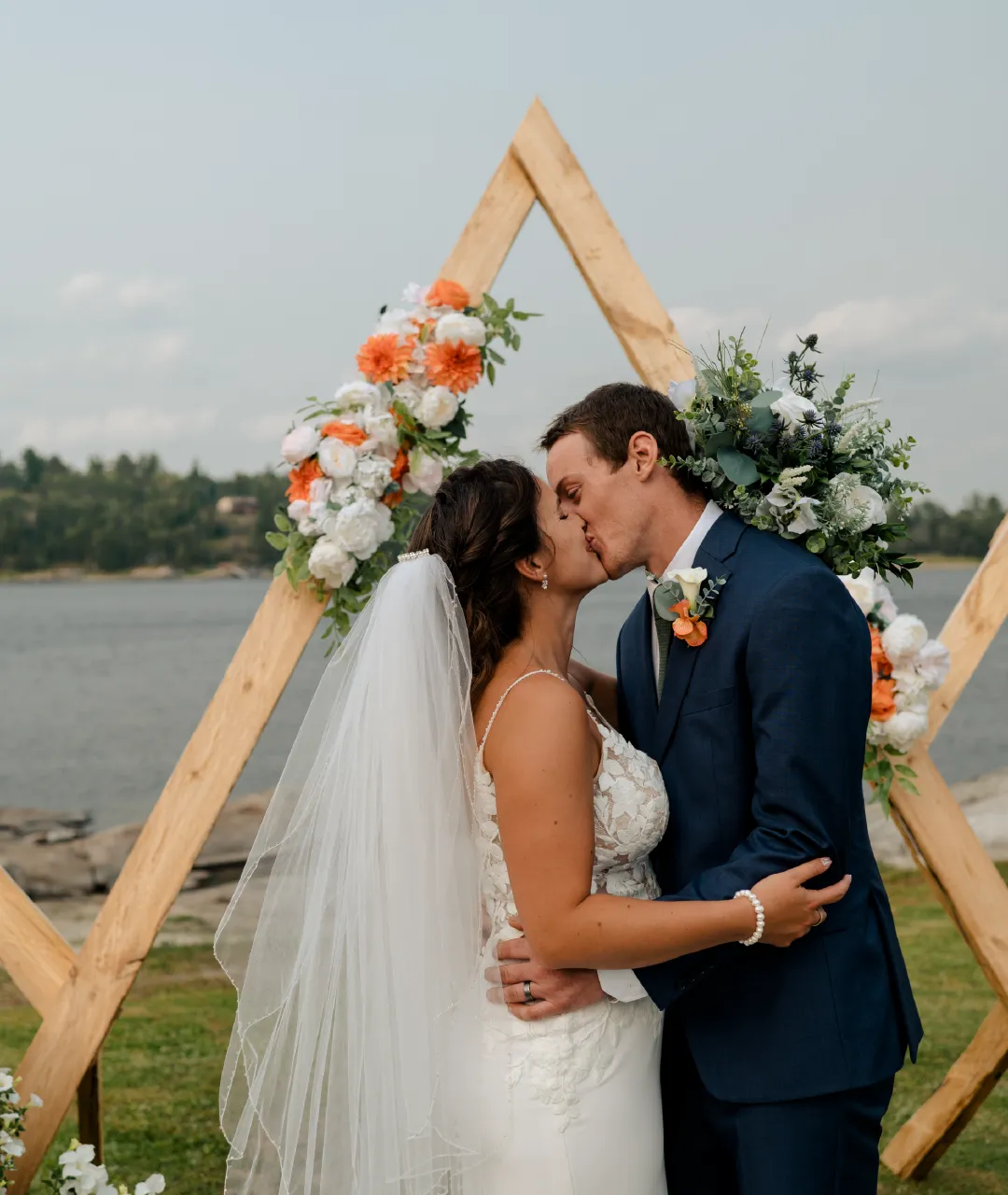 Wedding couple kissing in front of a wooden floral ceremony arch with the river in the background.