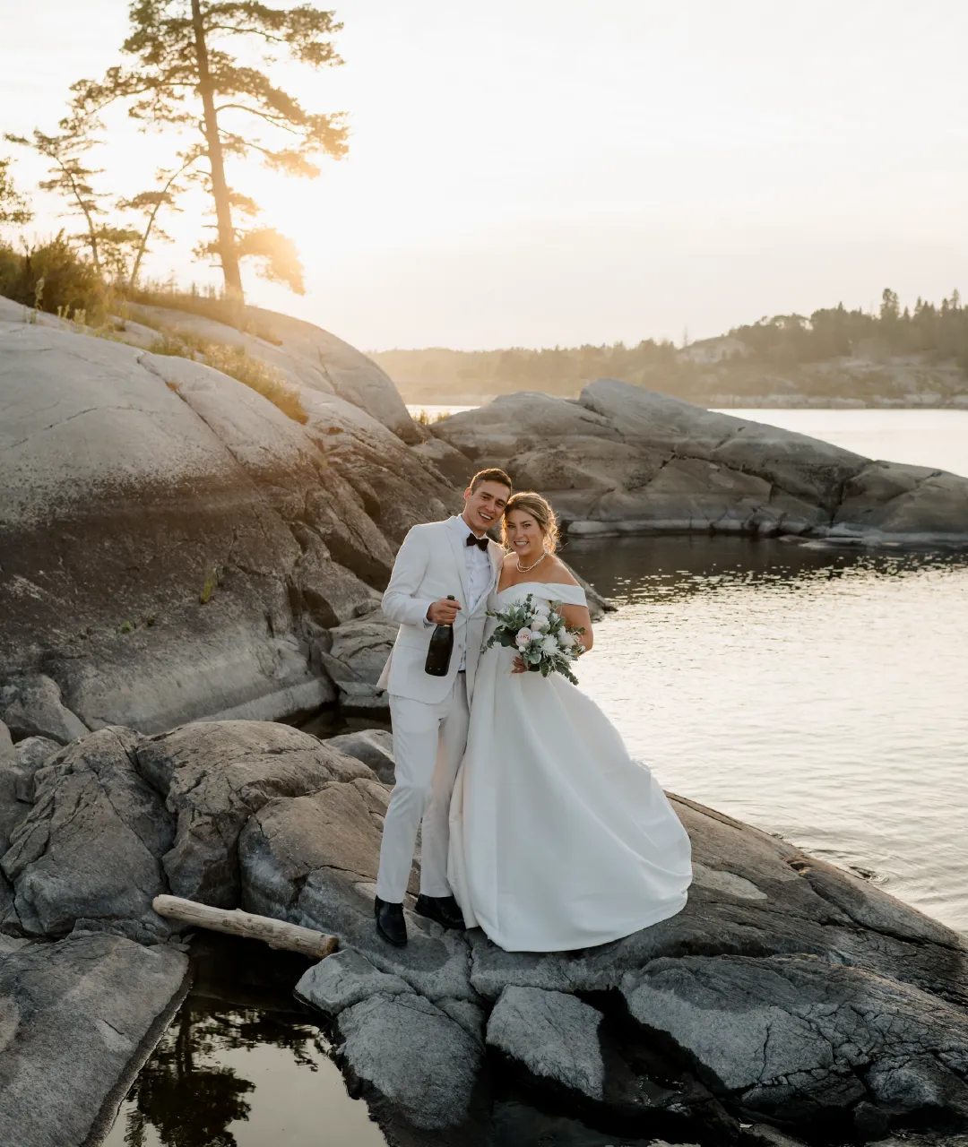 Wedding couple standing together on a rocky shoreline beside the water at sunset.