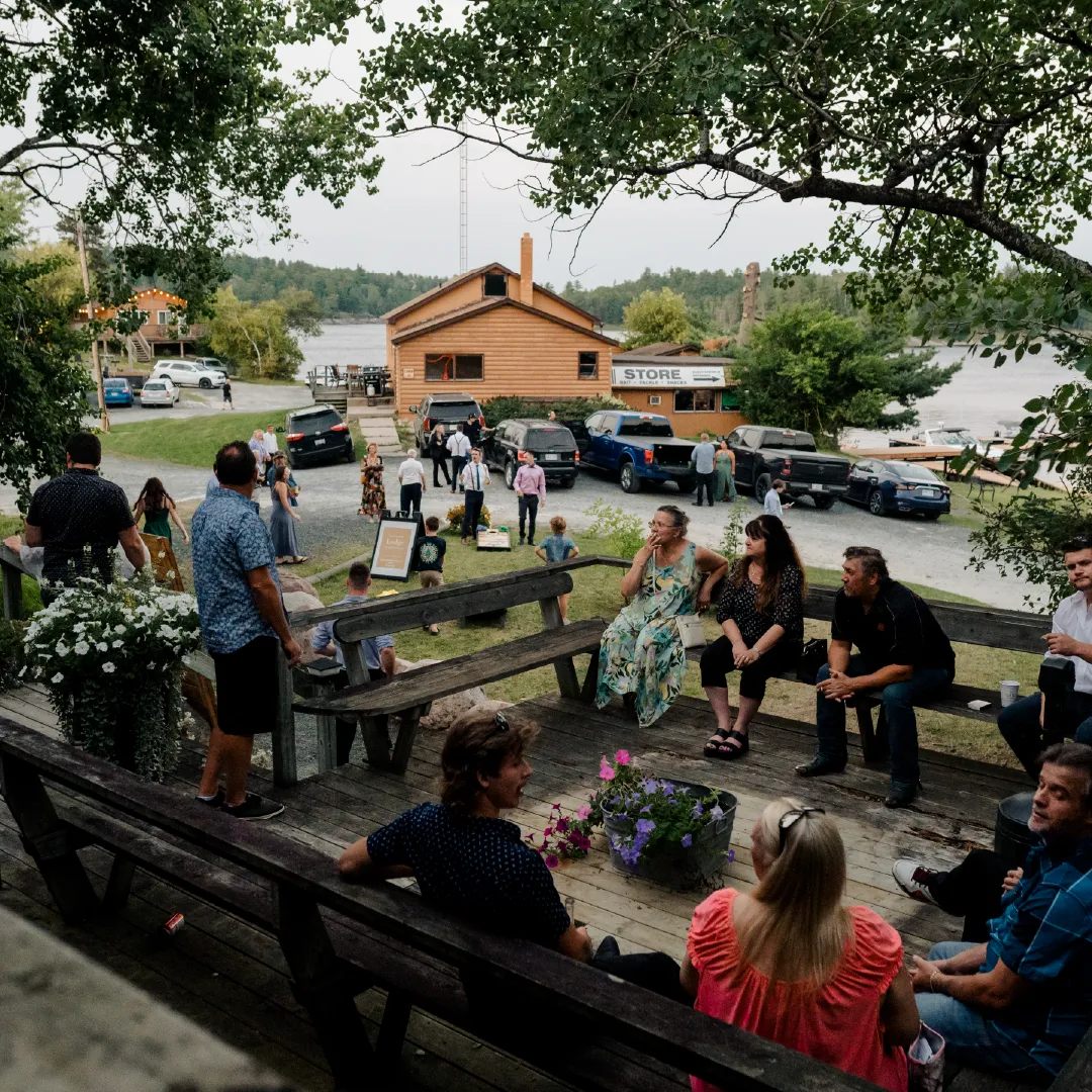 Wedding guests gathered on a wooden lodge deck overlooking the resort store, parked cars and river.