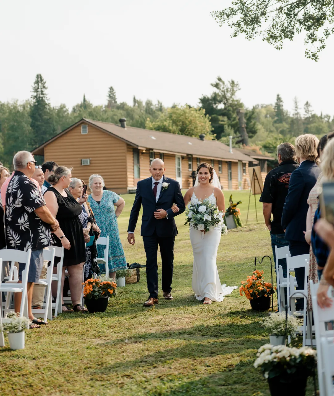 Bride walking down an outdoor ceremony aisle with a guest escort, white chairs and flower pots on the grass.