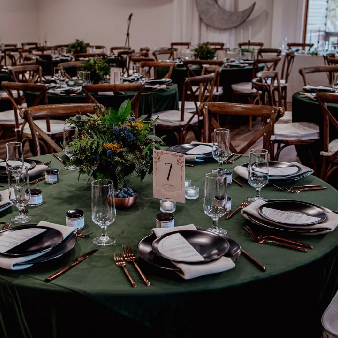 Wedding reception table set with green linens, dark plates, glassware, candles and a floral centrepiece.