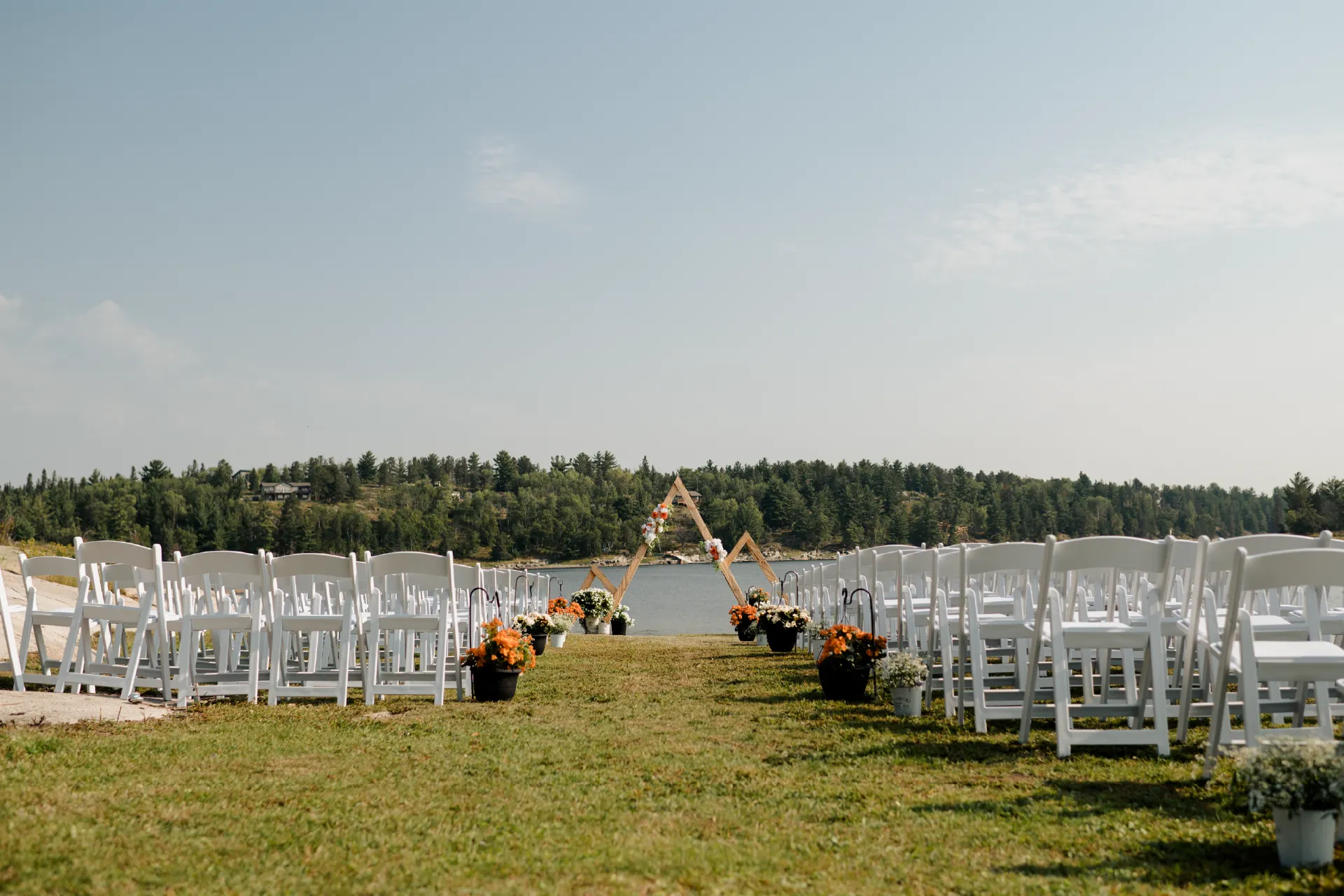 Outdoor wedding ceremony setup with white chairs, flower pots and a wooden arch facing the river.