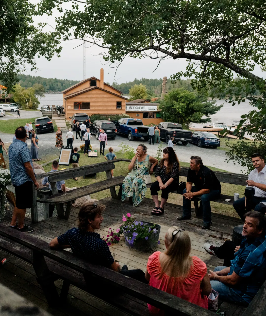 Guests sitting and standing on a wooden lodge deck overlooking the resort store, parked cars and river.