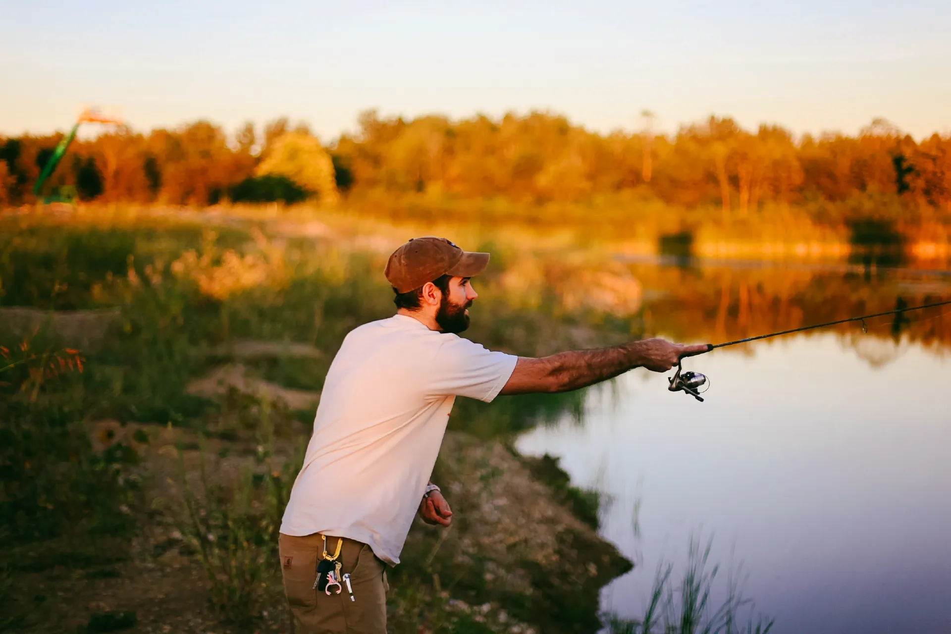 Person casting a fishing rod from the shoreline beside calm water at sunset.