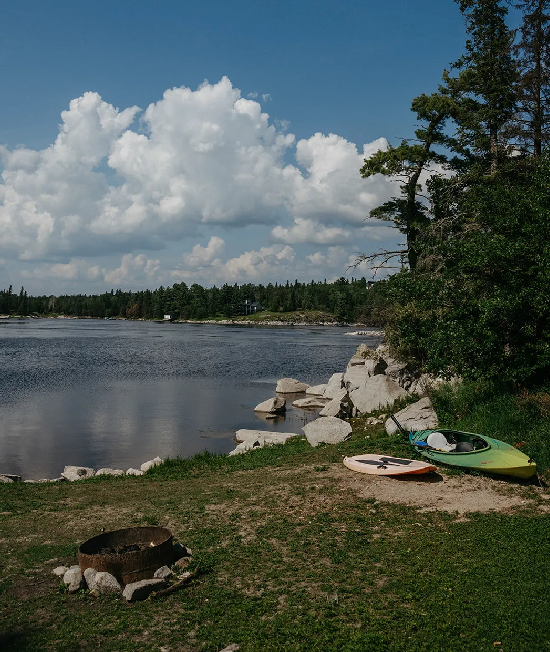 Rocky shoreline with a fire pit, kayak and paddleboard beside calm water under a cloudy sky.