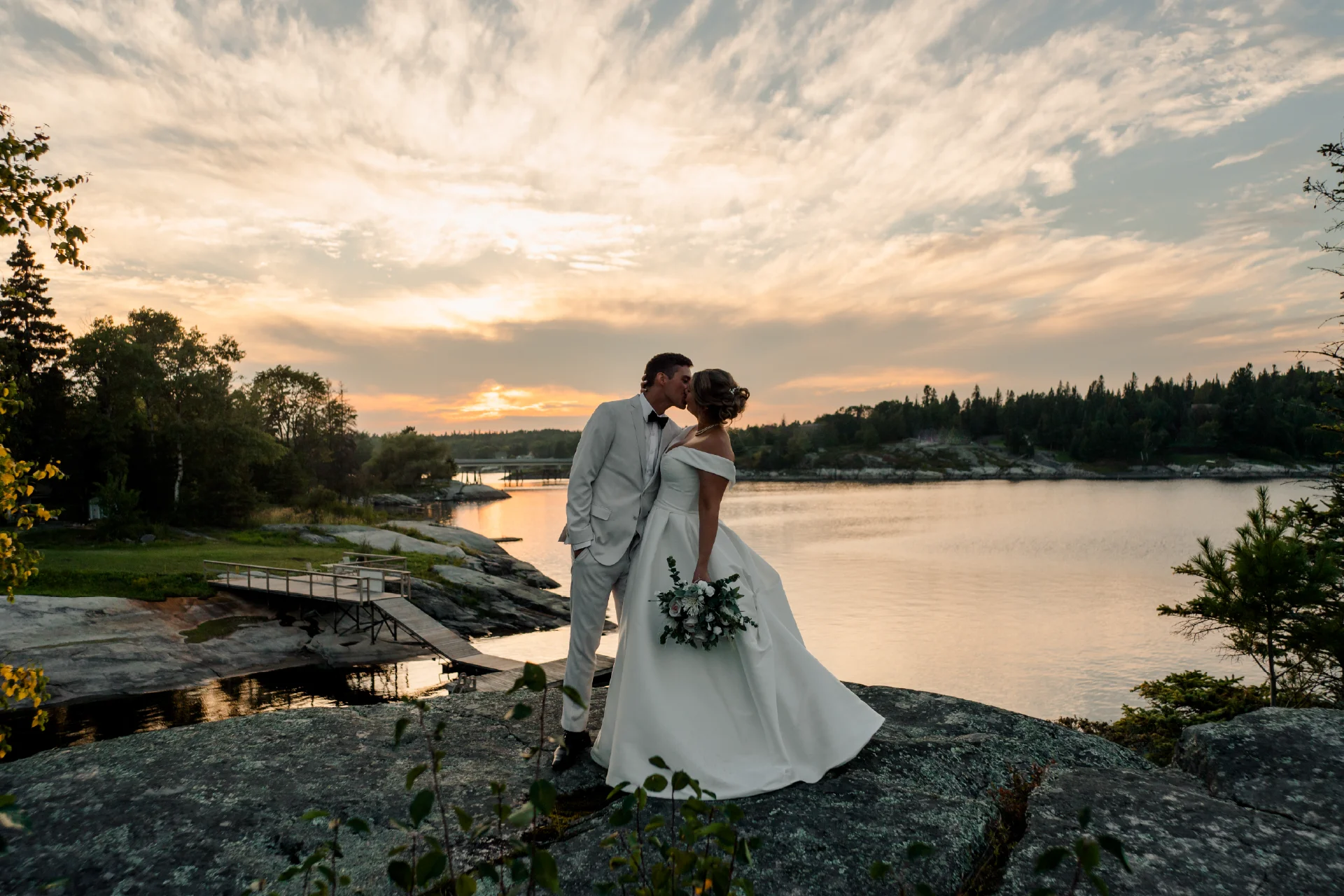 Wedding couple kissing on a rocky shoreline at sunset beside the river.