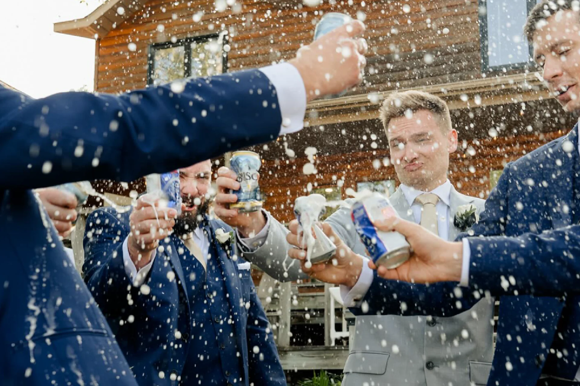 Wedding party members raising drinks outside a log lodge with spray in the air.