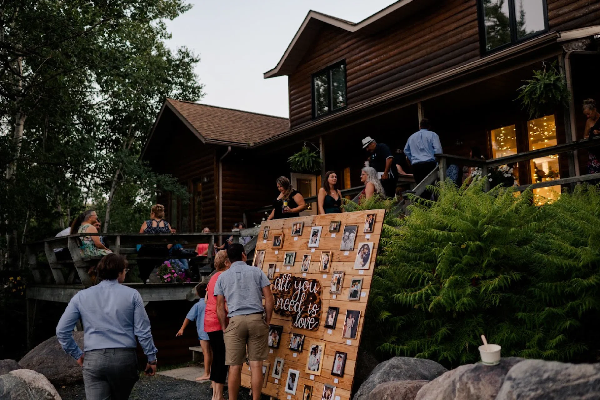 Wedding guests gathered outside a log lodge near a wooden photo display and fern-covered landscaping.