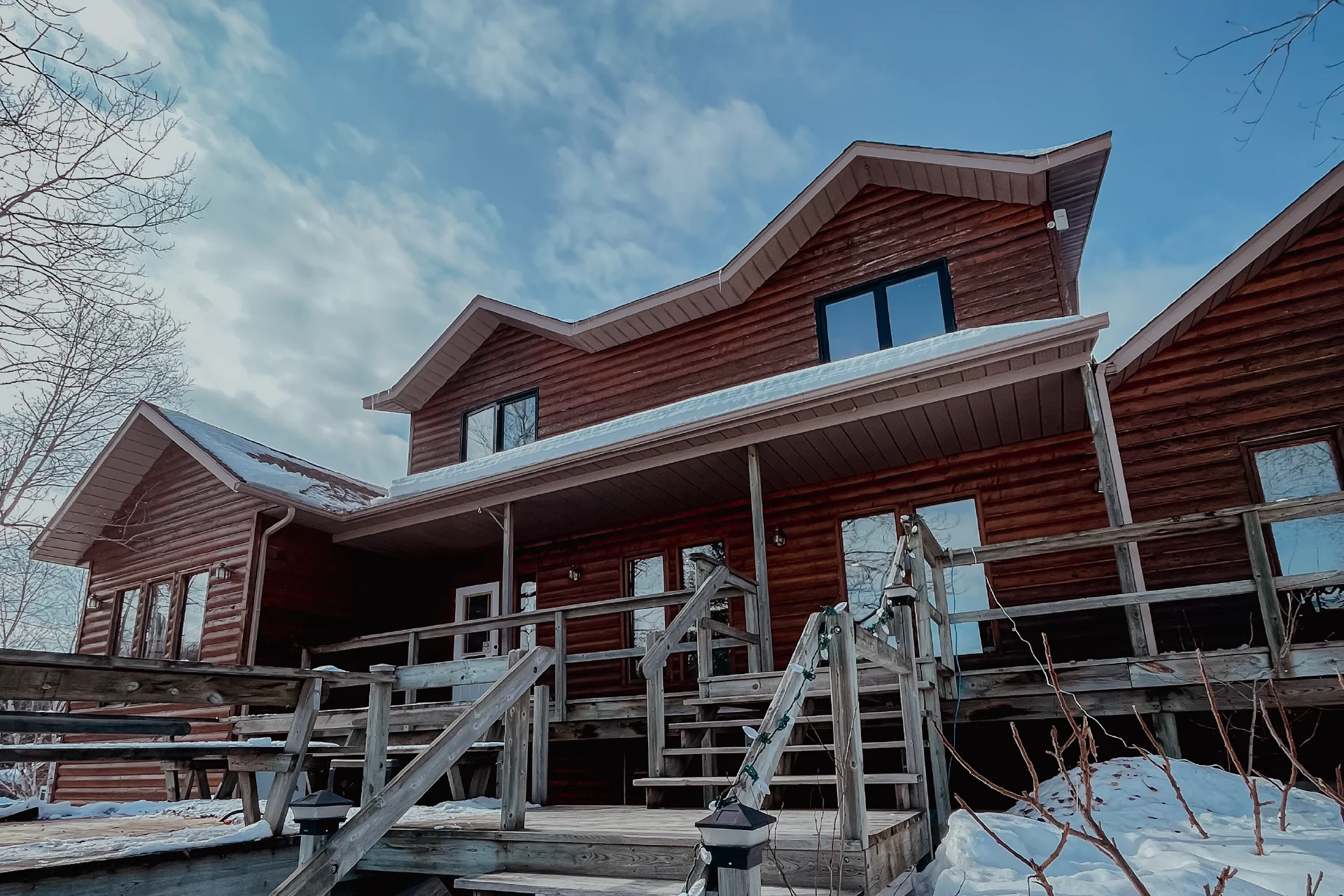 Exterior of a large log lodge with wooden stairs and railings surrounded by snow.