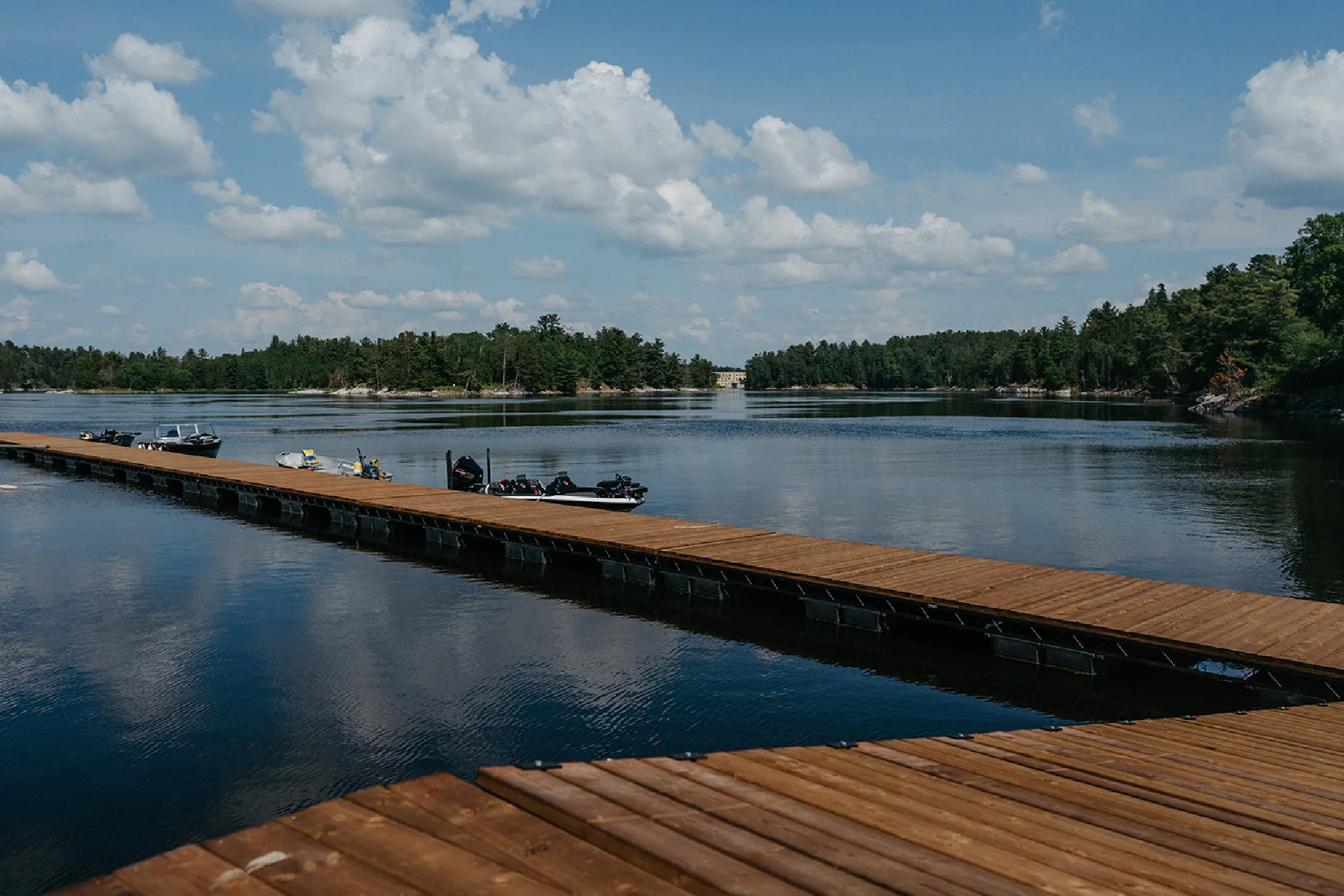 Long wooden dock with boats on calm water, surrounded by forested shoreline and cloudy sky.