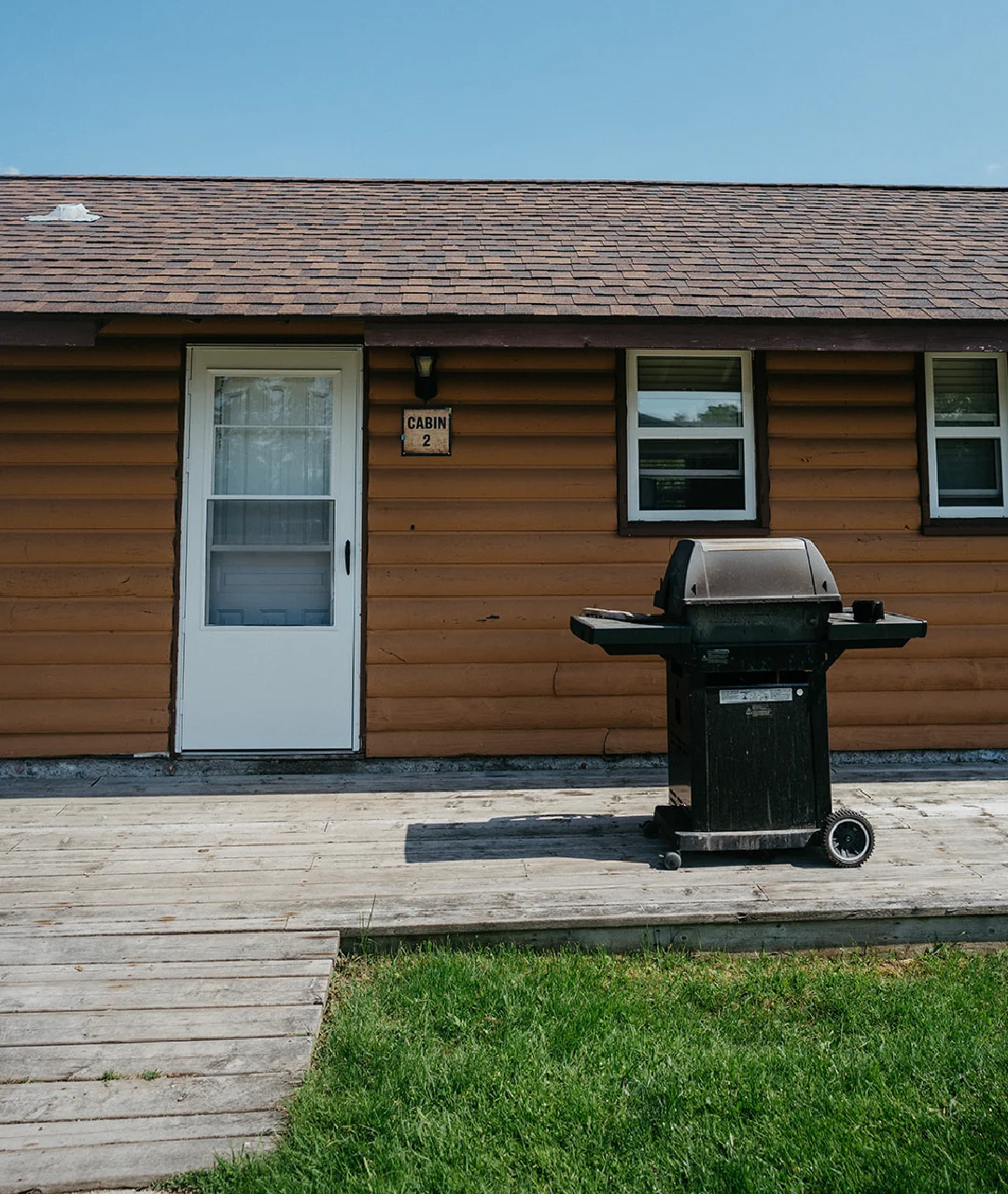 Exterior of Cabin 2 with a white door, wooden deck, barbecue grill and grass lawn.