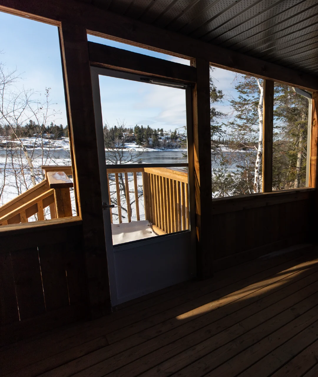 View from a covered cabin porch toward a snowy river shoreline through an open screen door.