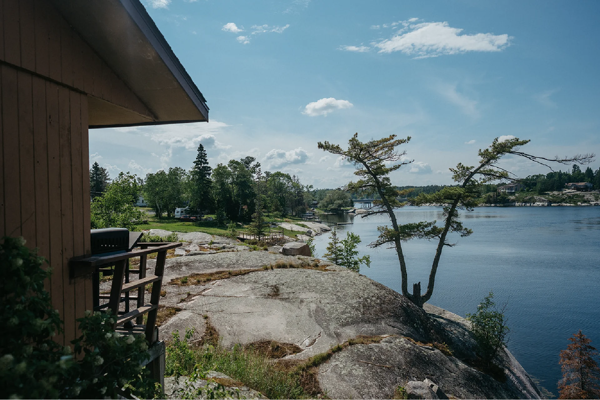 Cabin deck with a barbecue overlooking a rocky shoreline, trees and calm water.