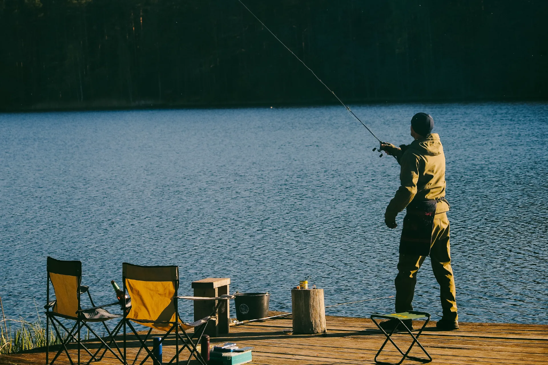 Person casting a fishing rod from a wooden dock with chairs and tackle beside them.