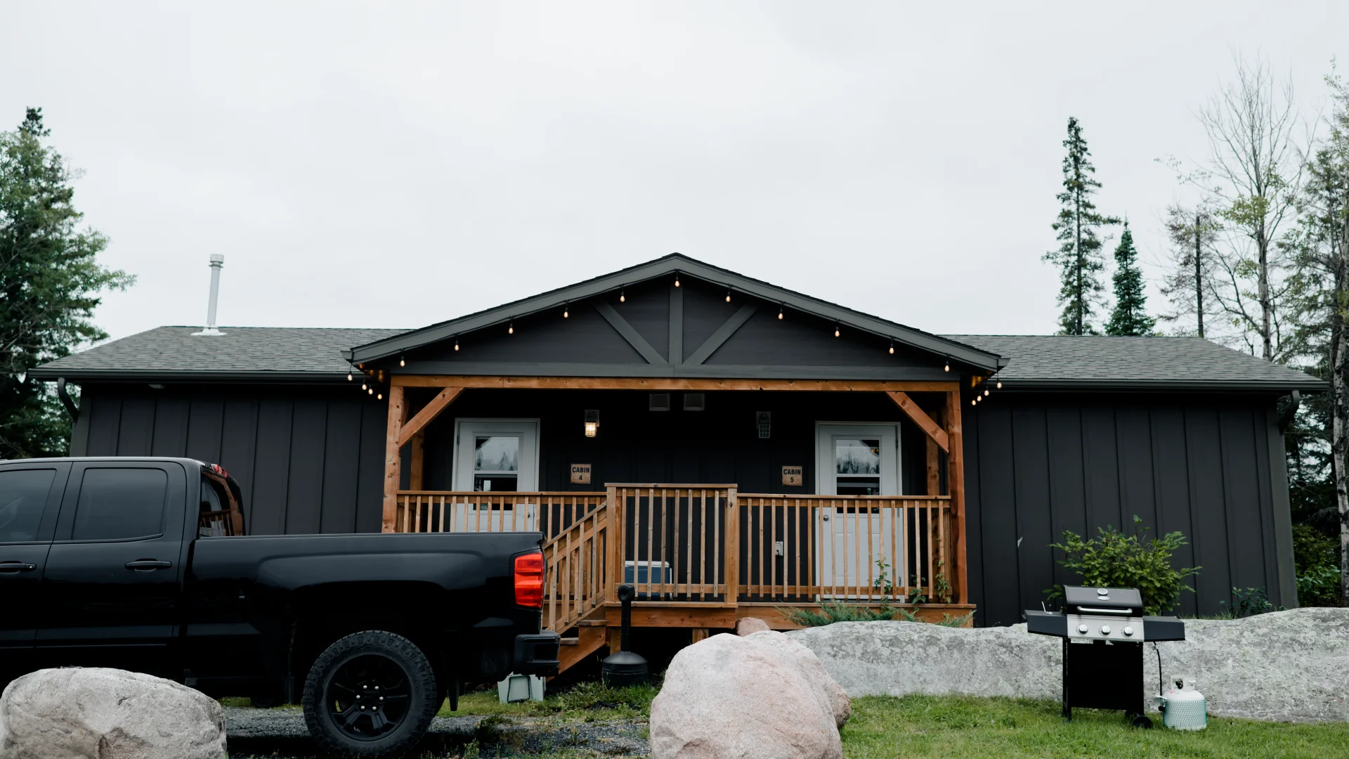 Exterior of the Beachfront Cabins with a covered wooden porch, string lights, large rocks, a barbecue, and a truck parked in front.