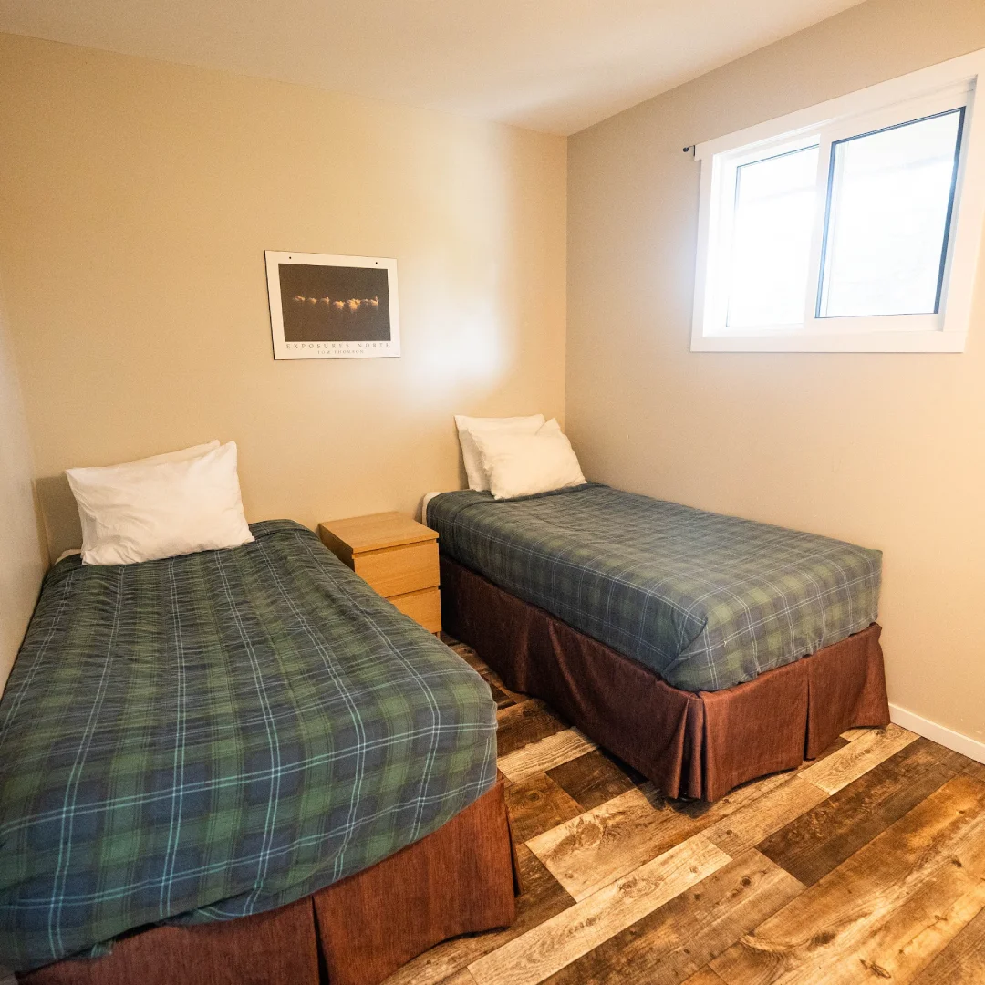 Simple suite bedroom with two twin beds, plaid blankets, white pillows, a nightstand, and a small window.