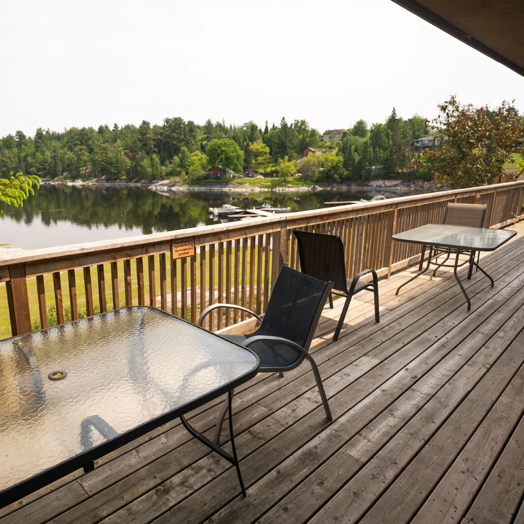 Wooden deck with patio tables and chairs overlooking the lake, docks, trees, and cabins on the opposite shore.